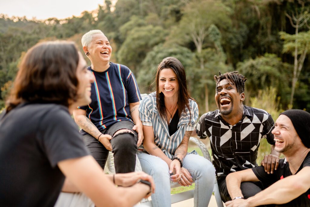 Group of diverse young friends laughing together outside in summer A group of adults sit outdoors smiling and laughing brightly