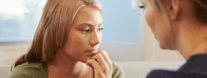 A closeup of a thoughtful teenage girl holding her hand to her face, as an adult woman gazes at her